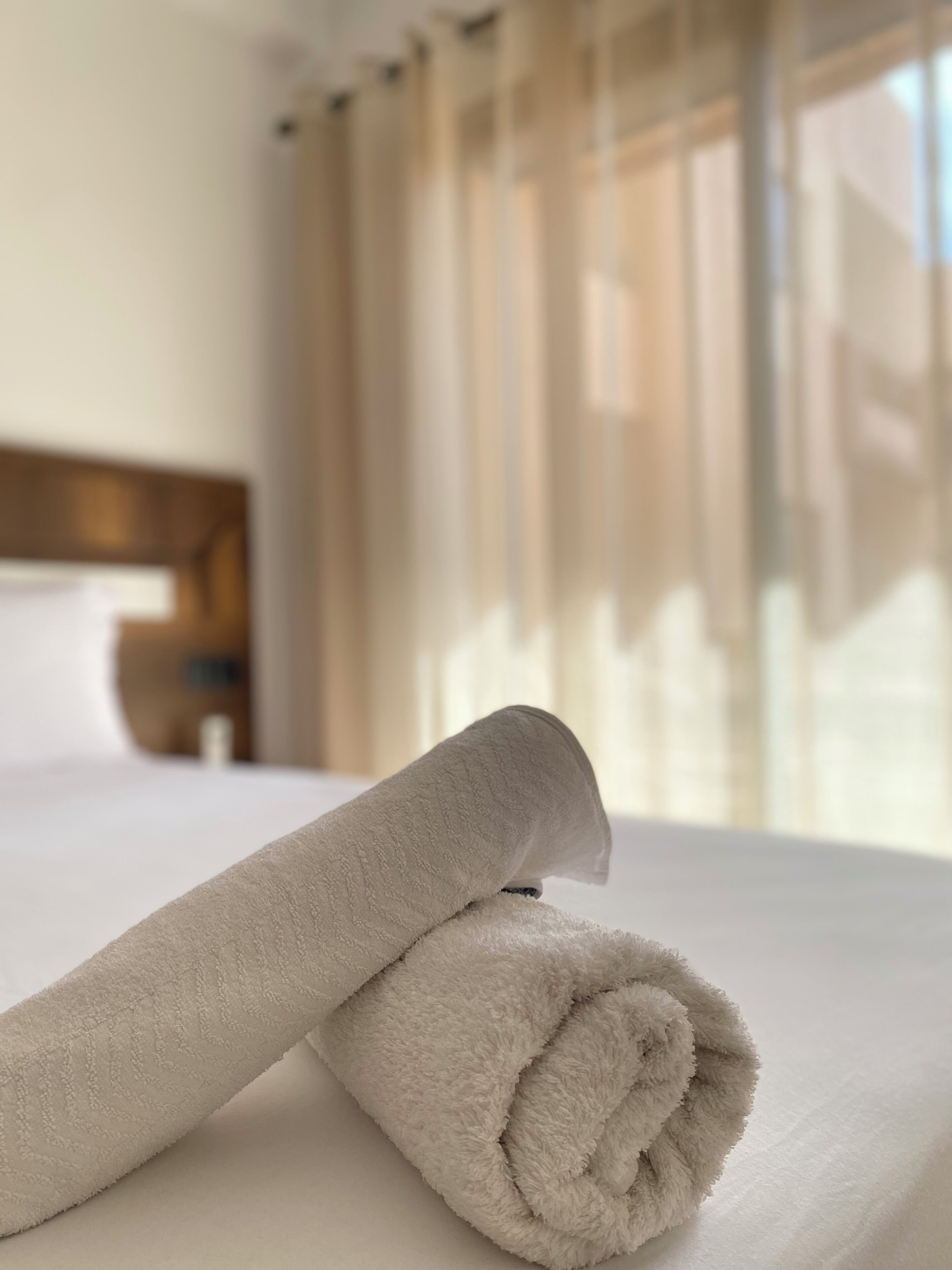 Een net opgemaakt bed met een opgerolde witte handdoek, een houten hoofdbord en lichte, doorschijnende gordijnen.