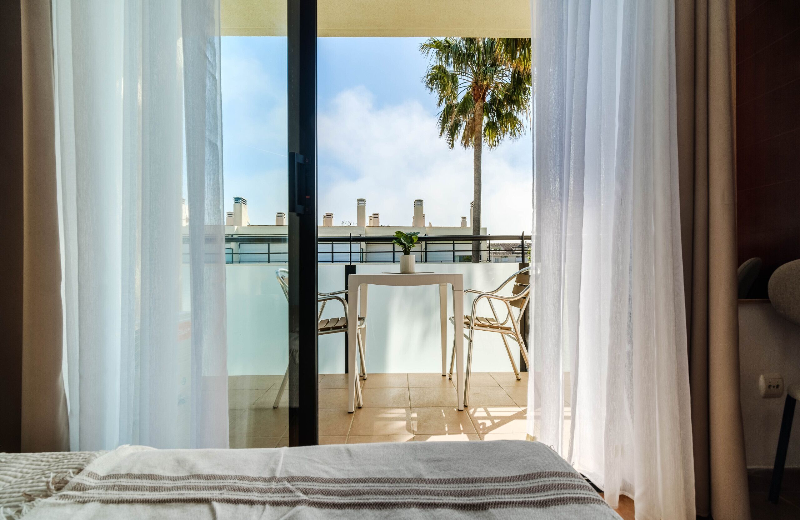 Un balcon avec une table et des chaises, un pot de fleurs et une vue sur des immeubles et des palmiers.