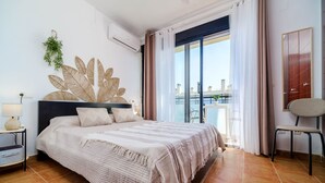 Ein Schlafzimmer mit Bett, einem Sessel, einem großen Fenster mit Vorhängen, einer Wandpflanze und einem Kleiderschrank.