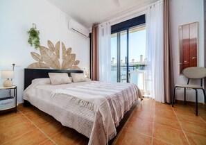 Ein Schlafzimmer mit Bett, einem Sessel, einem großen Fenster mit Vorhängen, einer Wandpflanze und einem Kleiderschrank.