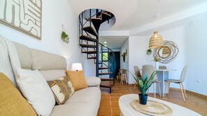 Ein modernes Wohnzimmer mit einer gebogenen Treppe, einer Couch mit gemusterten Kissen, einem runden Tisch mit geflochtener Tischmatte und einer hängenden Rattan-Leuchte.