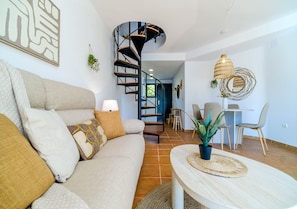 A modern living room with a curved staircase, a sofa with patterned cushions, a round table with a woven placemat, and a hanging rattan light fixture. - Santa Barbara Living Apartment Javea (Javea)
