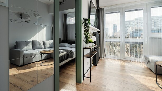 A modern living room with a grey sofa, a small table, a plant, and a view of the city through large windows. - RentPlanet - przy Parku Mirowskim (Warsaw)