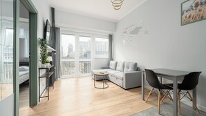 A modern living room with a grey sofa, a dining table with two chairs, a small round table, and a large window with a view of buildings. - RentPlanet - przy Parku Mirowskim (Warsaw)