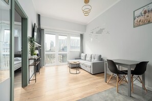 A modern living room with a grey sofa, a dining table with two chairs, a small round table, and a large window with a view of buildings. - RentPlanet - przy Parku Mirowskim (Warsaw)
