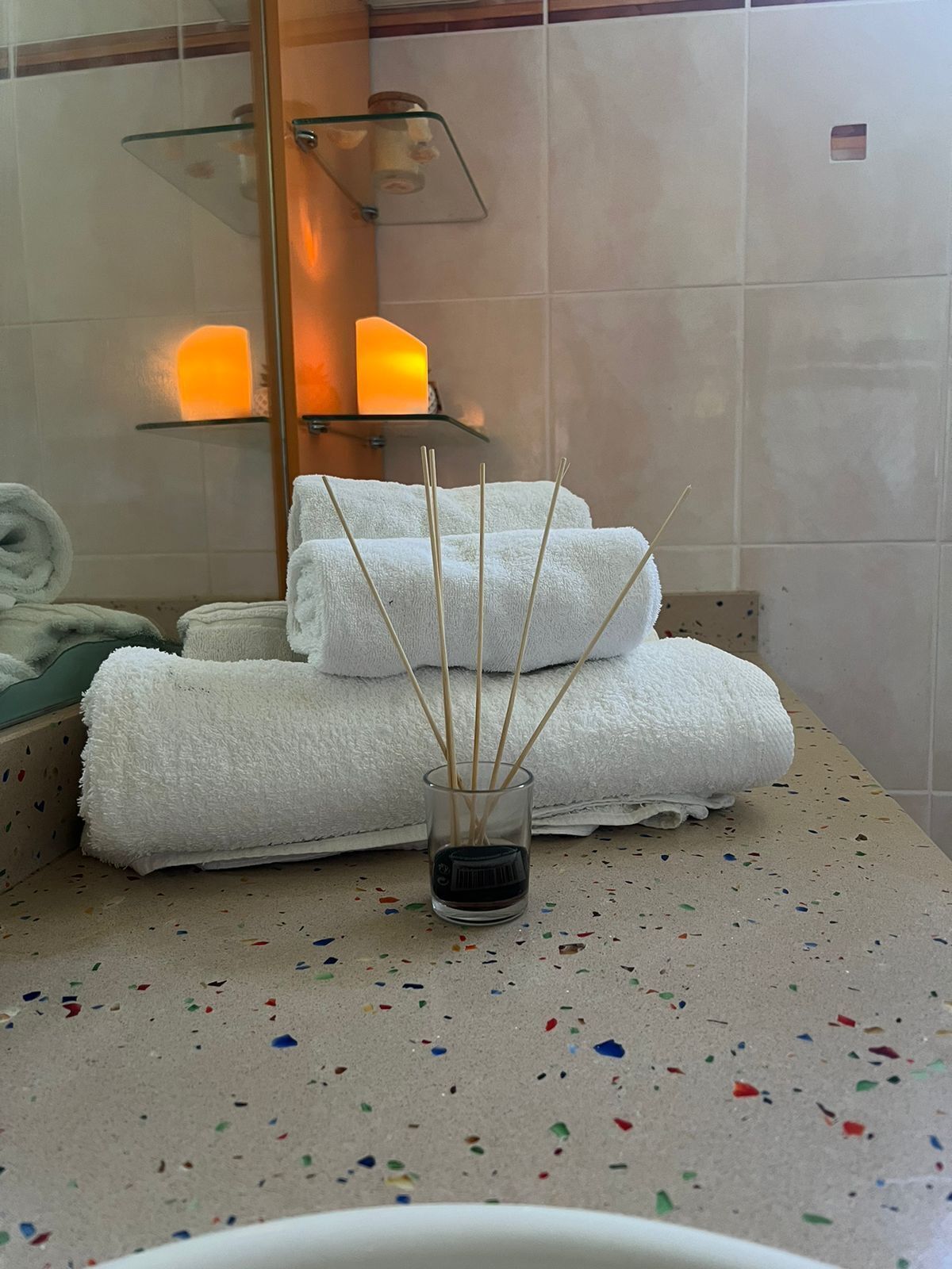 A bathroom with a glass shelf holding candles, a glass candle holder with incense sticks, and a towel on the counter.