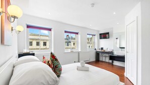 A bedroom with a bed, bedside tables, a desk, a chair, a TV, and a view of buildings through the windows. - Oriental Eight | By My Getaways (Brighton)