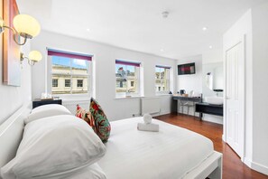Ein Schlafzimmer mit Bett, Nachttischen, Schreibtisch, Stuhl, Fernseher und Blick auf Gebäude durch die Fenster.