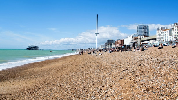 A pebbled beach with people, a pier, and buildings in the background. - Oriental Eight | By My Getaways (Brighton)