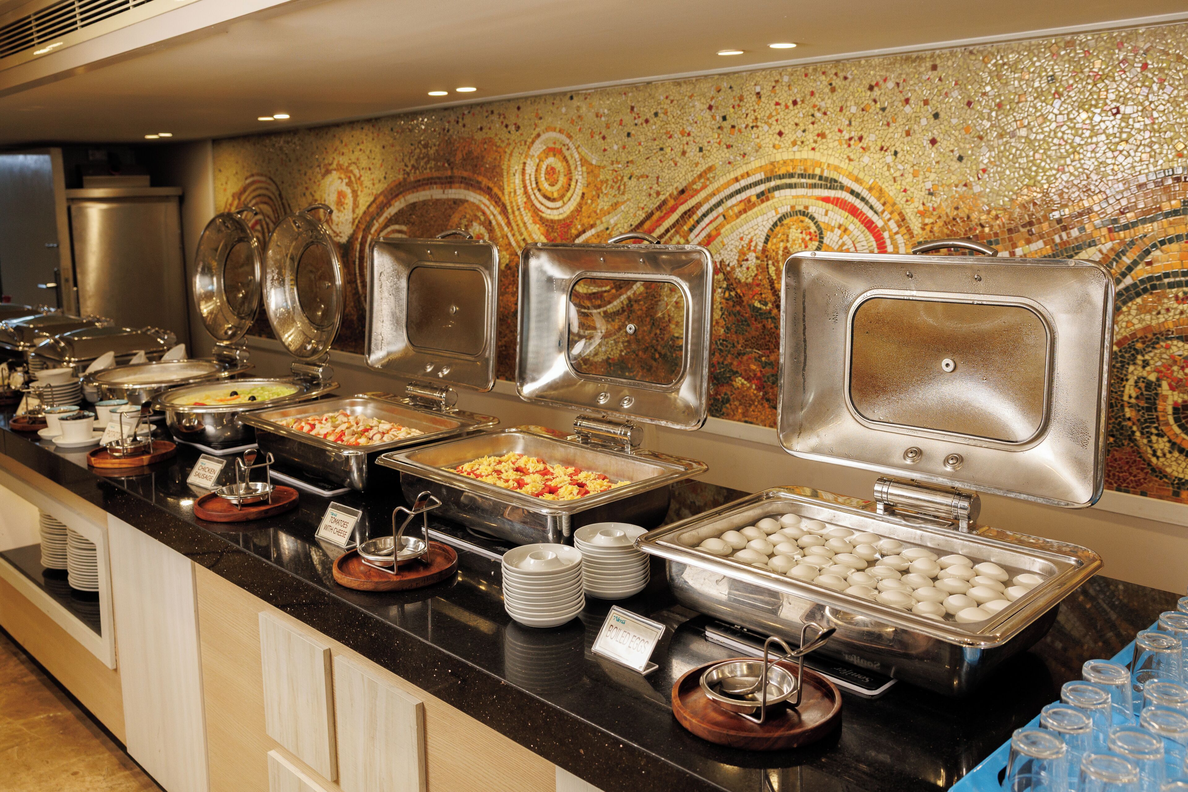 Buffet com diversas opções de comida, incluindo ovos, saladas e outros pratos, dispostos em um balcão com uma parede de mosaicos decorativos ao fundo.