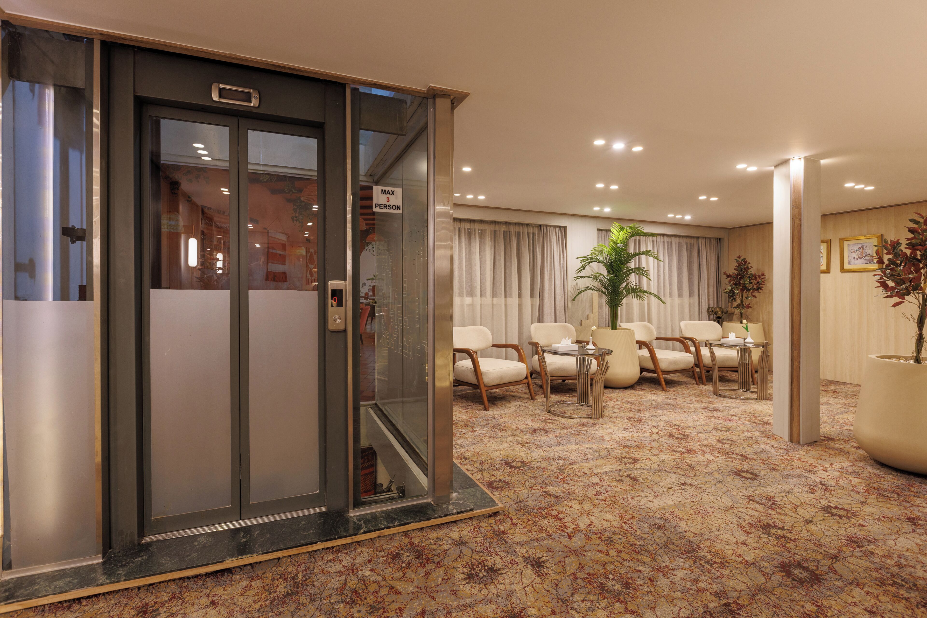 A hotel lobby with a glass elevator, a seating area with chairs and a table, and a carpeted floor.