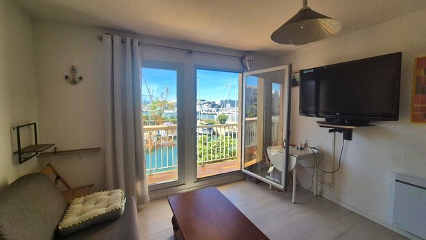 A room with a TV, a bench, a small table, a sofa, a window with a view of a marina, and a ceiling light. - Quai 12- 4Person apartment-marina view (Canet-en-Roussillon)