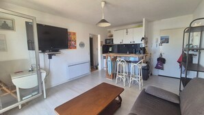 A living room with a TV, a wooden bench, a sofa, and a kitchen area with a bar stool. - Quai 12- 4Person apartment-marina view (Canet-en-Roussillon)