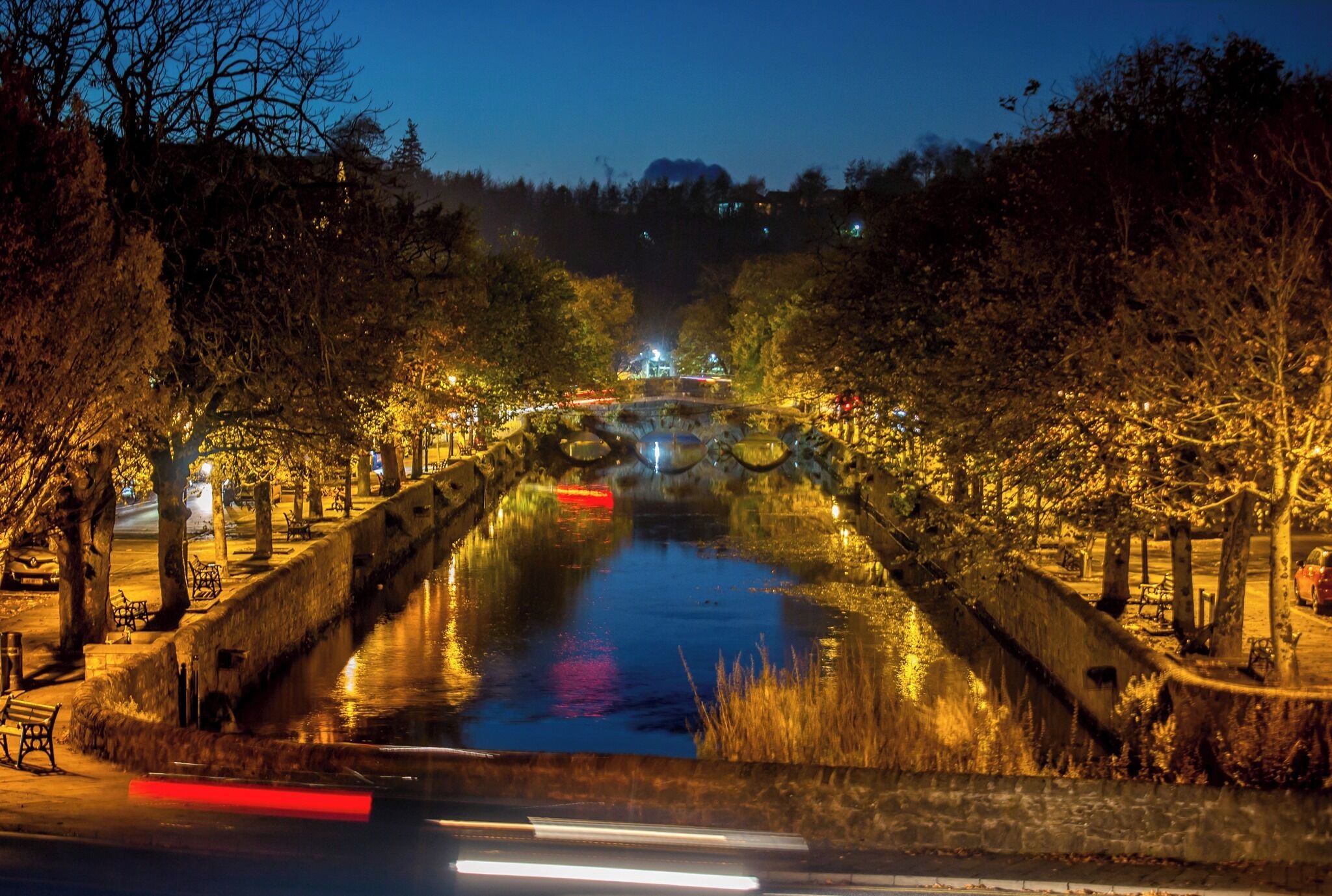 Un puente de piedra sobre un canal, flanqueado por árboles, iluminado por farolas.