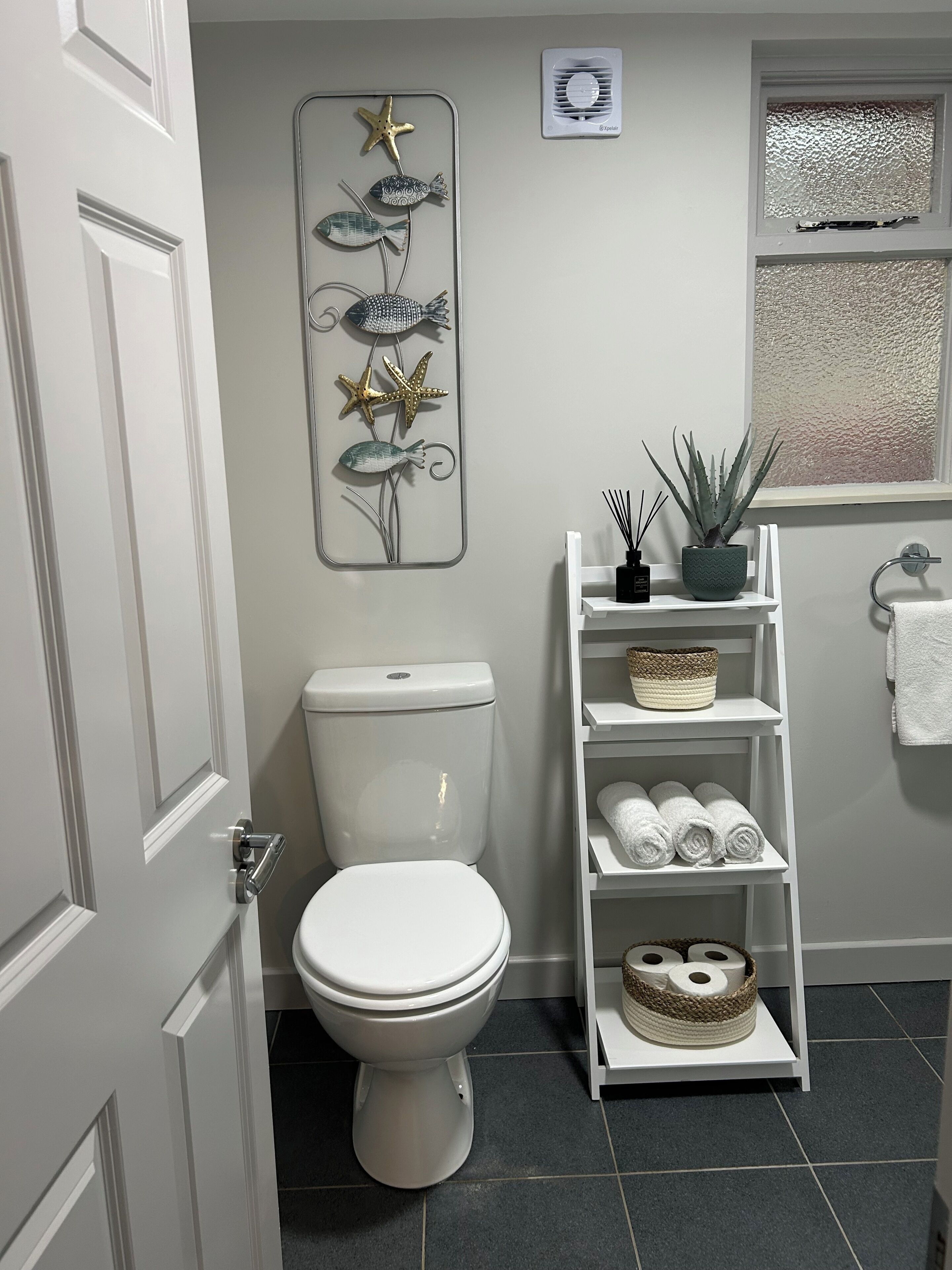 A bathroom with a white toilet, a decorative wall art piece, a potted plant, a towel rack, and a shelf with towels and a basket.