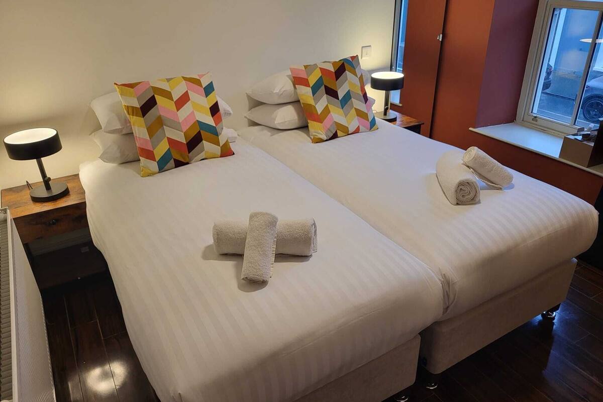 Hotelkamer met twee bedden, elk met een witte spreidek en kleurrijke kussens, een nachtkastje met een lamp en een raam met uitzicht.