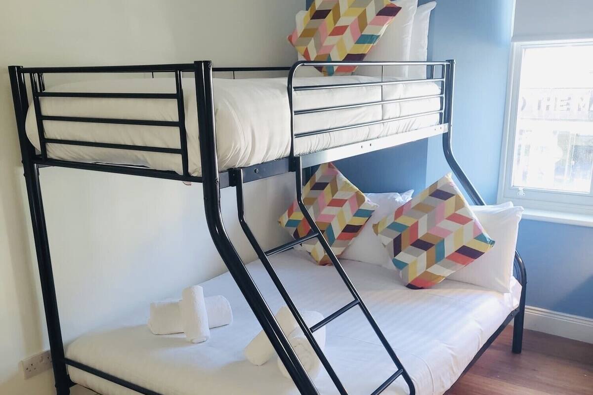 Een stapelbed met witte beddengoed en kleurrijke geometrische kussens, houten vloeren en een raam met daglicht.