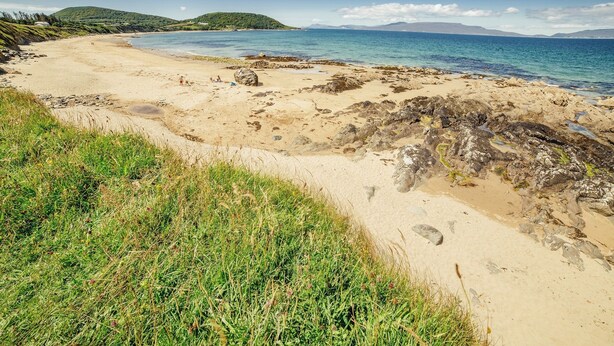 Ein Strand mit klarem Wasser, Felsen und grünen Hügeln im Hintergrund.