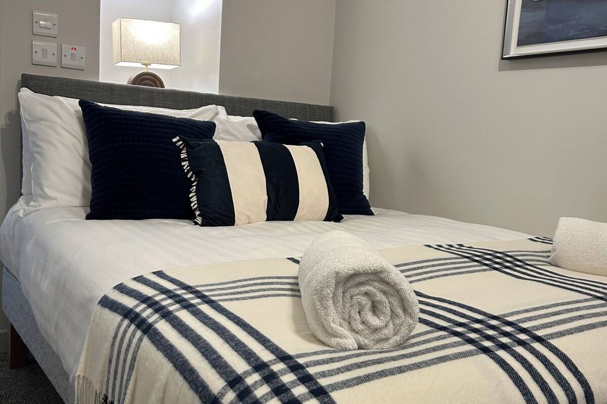 Een net opgemaakt bed met een geruit plaid, een opgerolde handdoek en meerdere kussens.