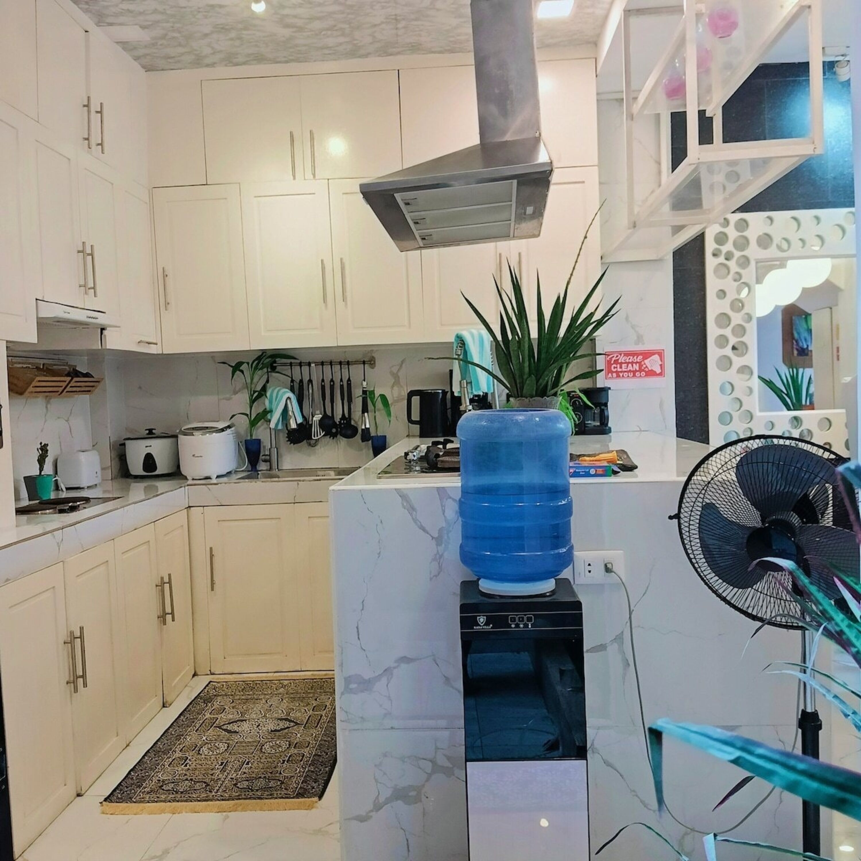 A modern kitchen with white cabinets, a large water dispenser, a fan, and a small rug.