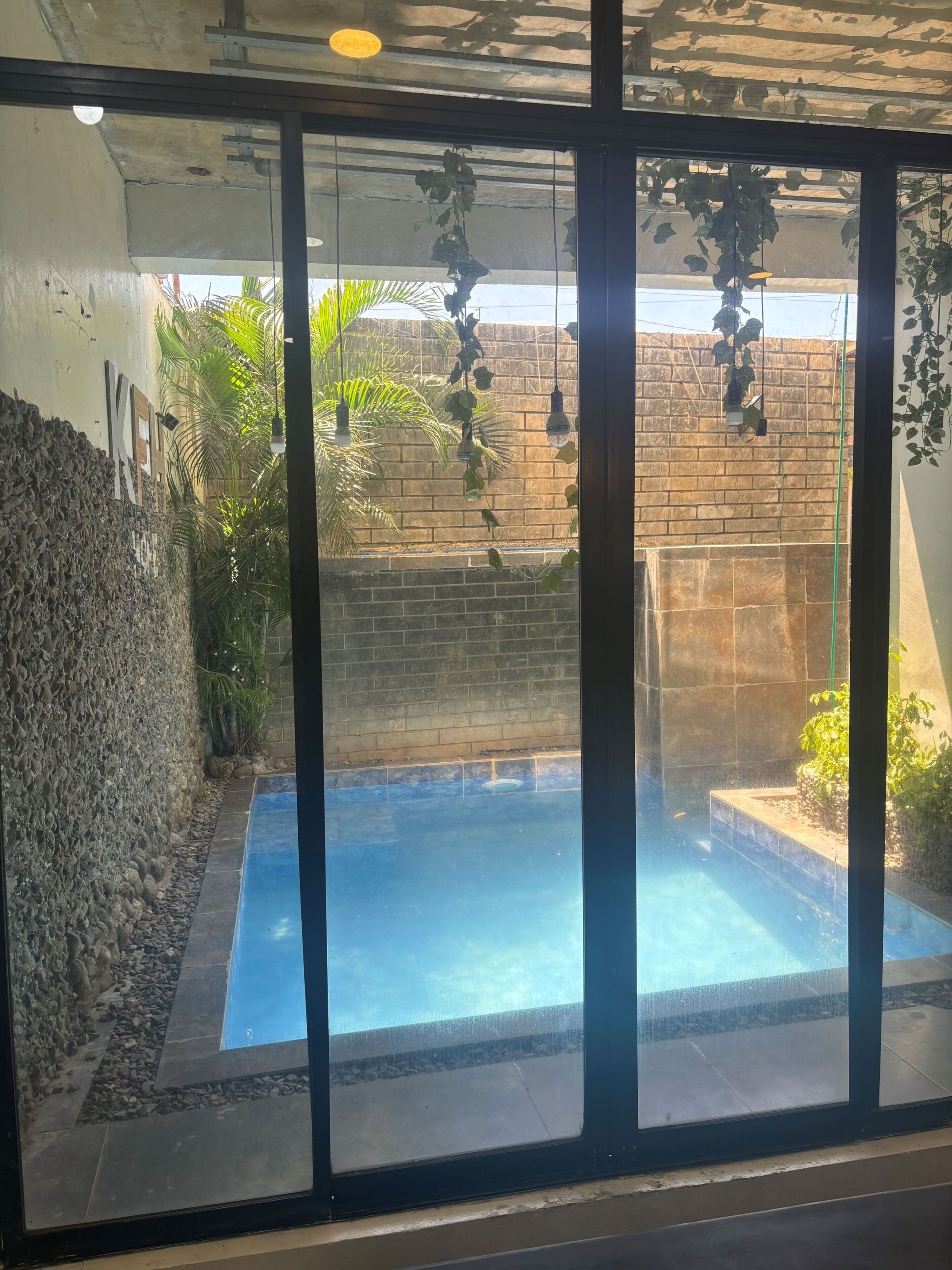 A swimming pool with a stone wall and plants, viewed through a glass door.