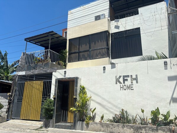 A modern building with a 'KFH HOME' sign, a gated entrance, and a balcony with plants.