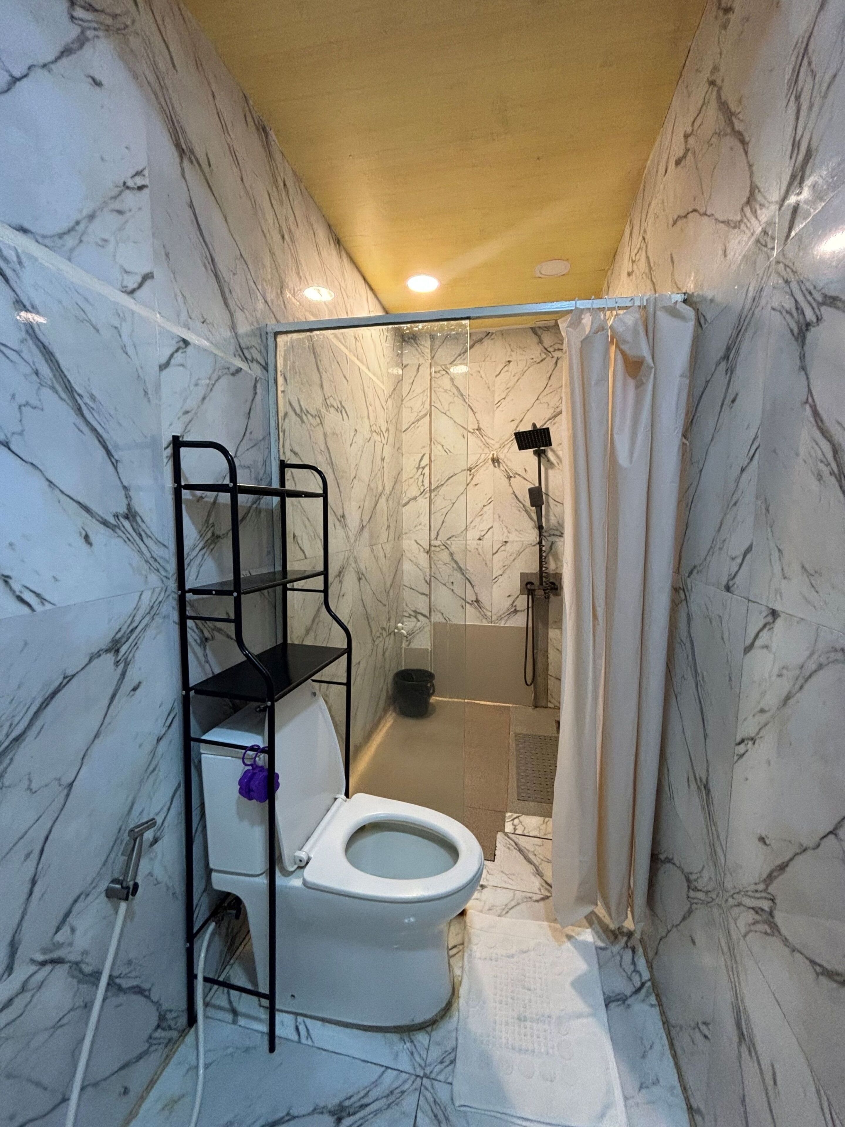 A bathroom with a toilet, shower, marble walls, and a shelf unit.