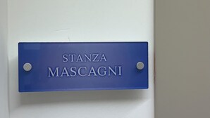 Een blauw bord met de tekst 'STANZA MASCAGNI' bevestigd aan een witte muur.