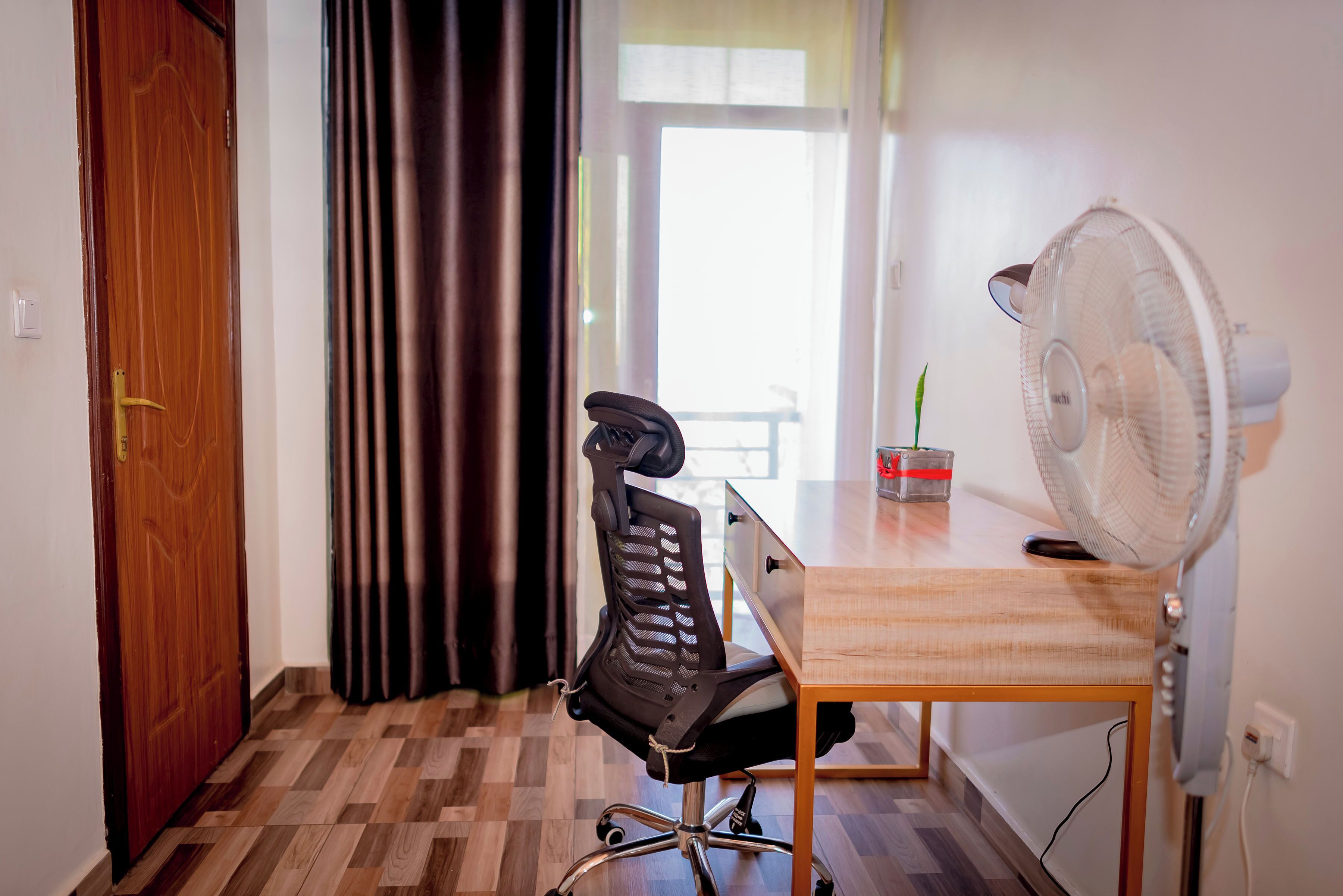 Une pièce avec une porte en bois, un fauteuil de bureau noir, un bureau en bois et un ventilateur sur pied.