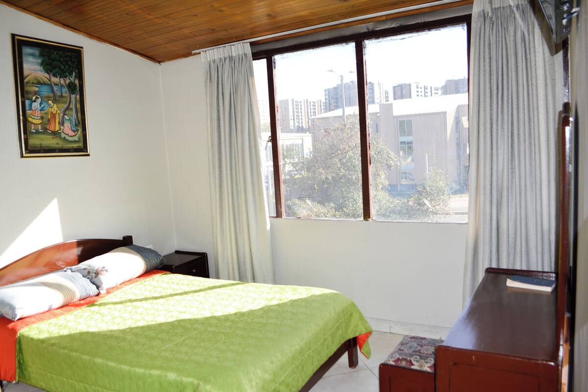 A bedroom with a bed, a nightstand, a dresser, a window with curtains, and a painting on the wall.