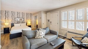A hotel room with a bed, a sofa, a chair, a TV, and a window with blinds. - Modern Rustic North Creek | Pool, Hot Tub, Tennis (The Blue Mountains)