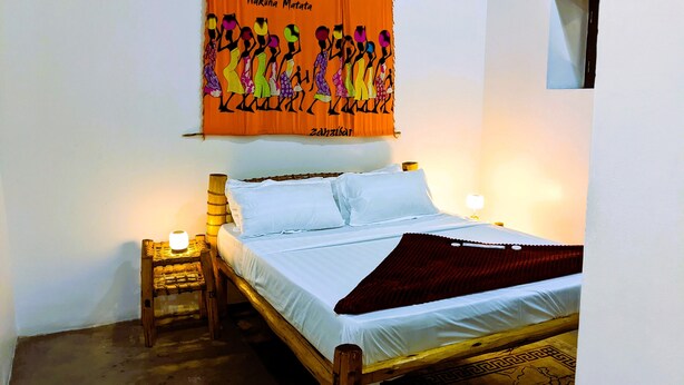 Ein Bett mit weißen Bettwäsche, einem hölzernen Kopfteil und einer roten Tagesdecke. Ein kleiner Nachttisch mit Lampe und ein Wandbild befinden sich darüber.