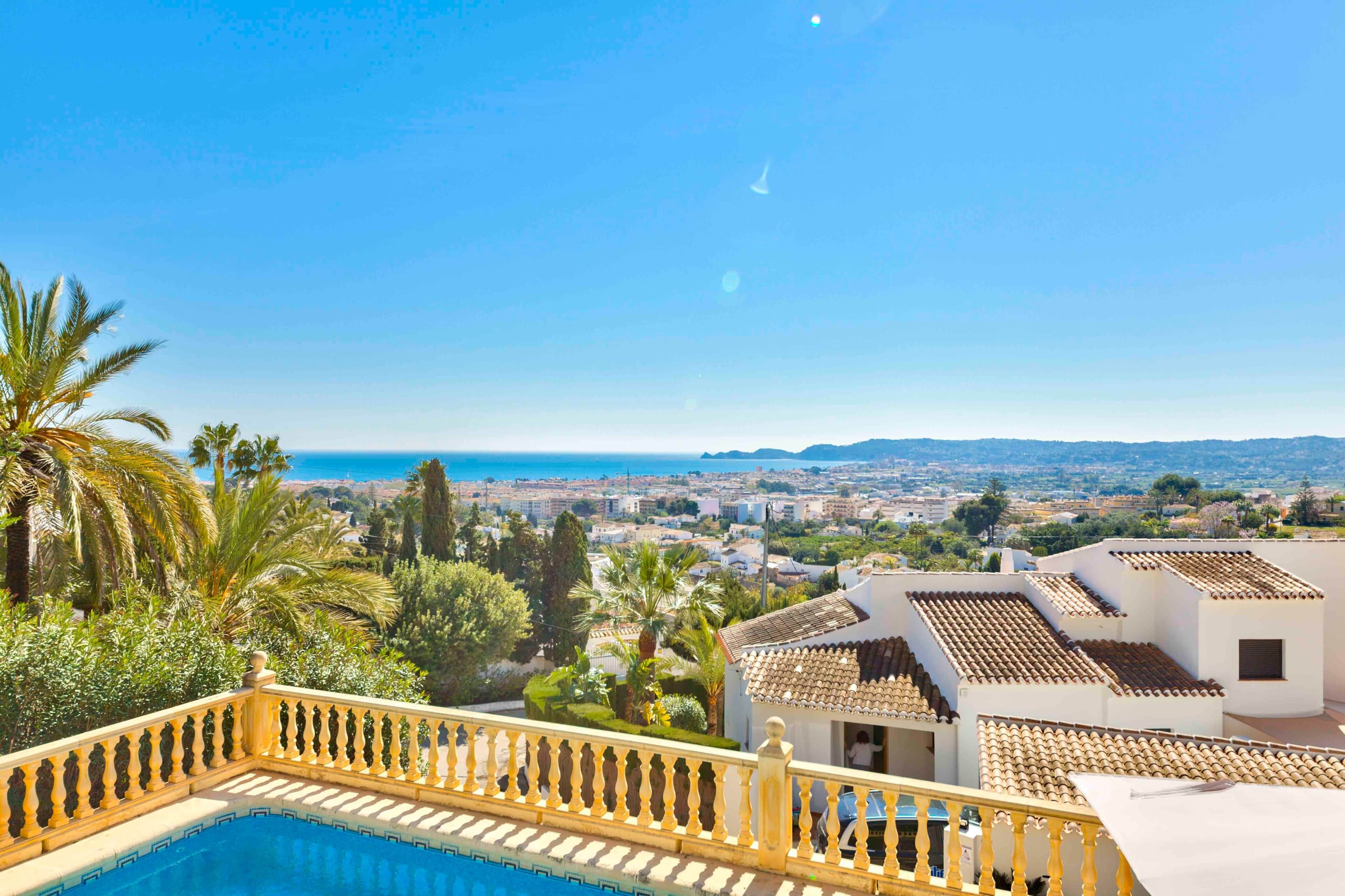 Une piscine bordée d’une balustrade jaune, avec vue sur la mer, et des bâtiments aux toits de tuiles.