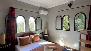 Una camera da letto con un letto, una poltrona blu, un comodino in legno e una finestra vista alberi.