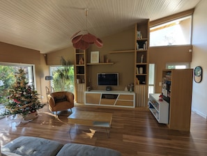 Ein modernes Wohnzimmer mit Holzboden, einem großen Fenster, einem Weihnachtsbaum, einer Couch, einem Couchtisch und einem Fernseher auf einem Holzständer.