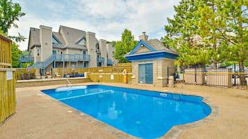 A swimming pool with a wooden deck, a small building with a blue roof, and a multi-story building in the background.
