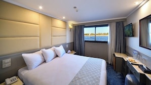A hotel room with a large bed, a desk, a chair, a TV, and a window with a view of the water. - MS Nile Treasure Cruise (Luxor)