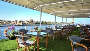 An outdoor seating area with tables and chairs, a view of the river, and buildings in the background. - MS Nile Treasure Cruise (Luxor)
