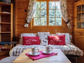 A cozy cabin room with a wooden interior, a large window with curtains, a bed with patterned pillows, and a table with two cups and saucers.