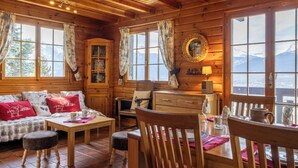 Ein gemütliches Blockhaus-Interieur mit einem Holzespeisetisch, Stühlen, einer Couch mit roten Kissen, einem Schrank und großen Fenstern mit Blick auf die Berge.