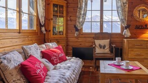 Ein gemütliches Blockhaus-Zimmer mit einer Couch, einem Sessel, einem Tisch und Blick auf verschneite Berge.