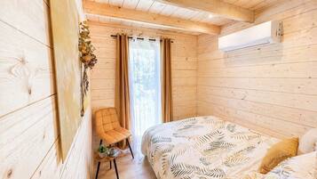 Een houten kamer met een bed, een stoel, een klein tafeltje met een kopje en een plant, en een raam met gordijnen.