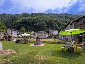 Un área de estar al aire libre con encanto, con mesas y sillas de piedra, una rueda hidráulica y vistas a edificios tradicionales y una colina boscosa.