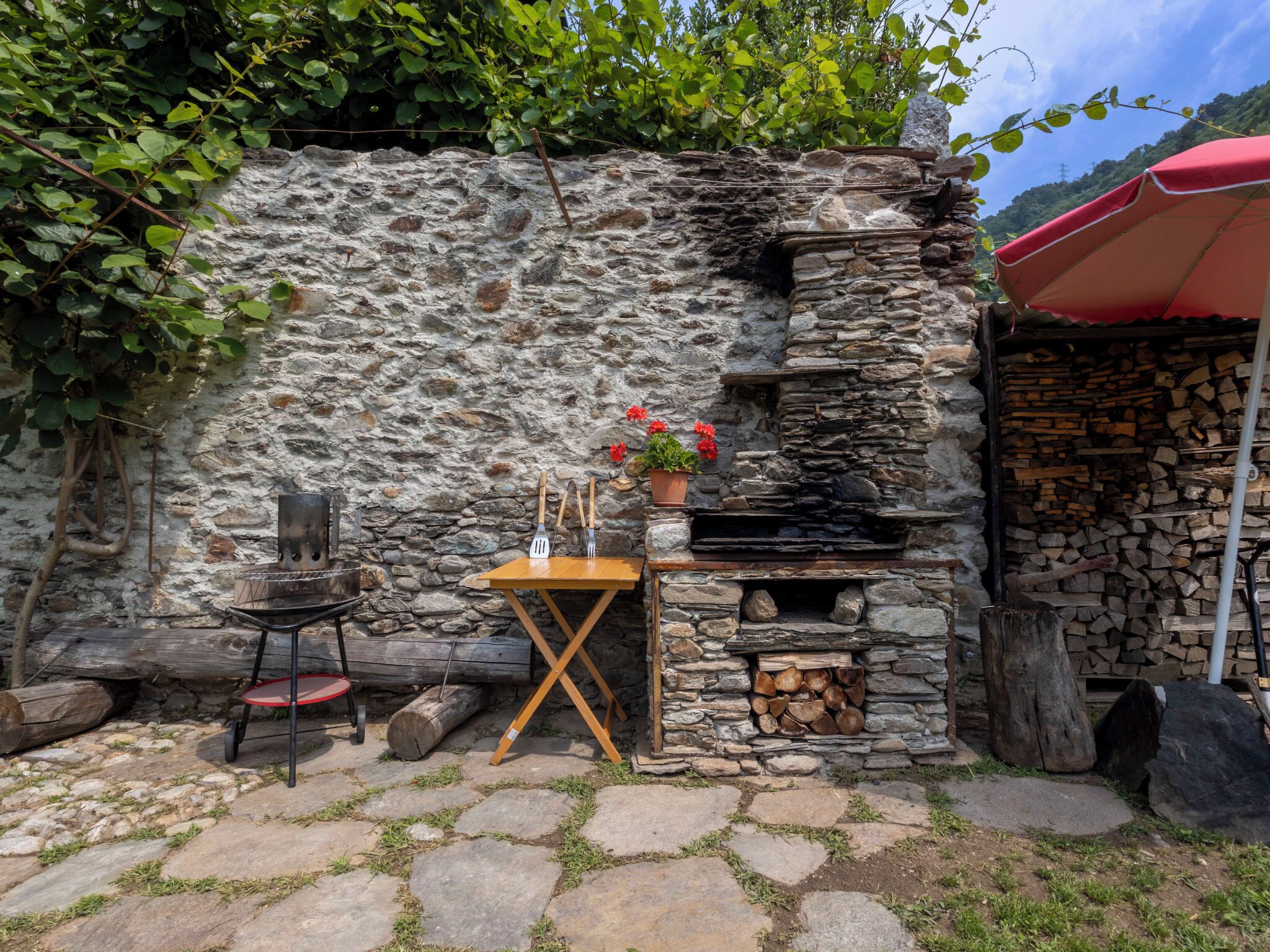 A stone wall with a small table and chairs, a grill, and a stack of firewood.