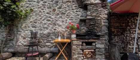 A stone wall with a small table and chairs, a grill, and a stack of firewood.