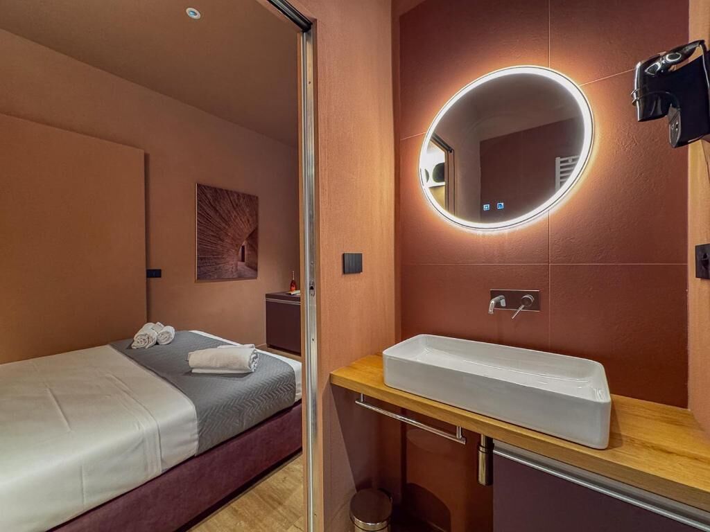Una habitación de hotel con cama, un espejo redondo con iluminación, un lavamanos y un pequeño cubo de basura.