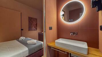 Una habitación de hotel con cama, un espejo redondo con iluminación, un lavamanos y un pequeño cubo de basura.