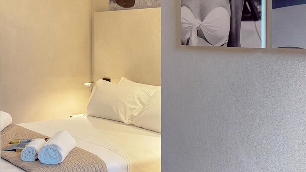 A hotel room with a bed, pillows, a rolled towel, and artwork on the wall. - Blu Rooms - Room to Rest (Civitanova Marche)