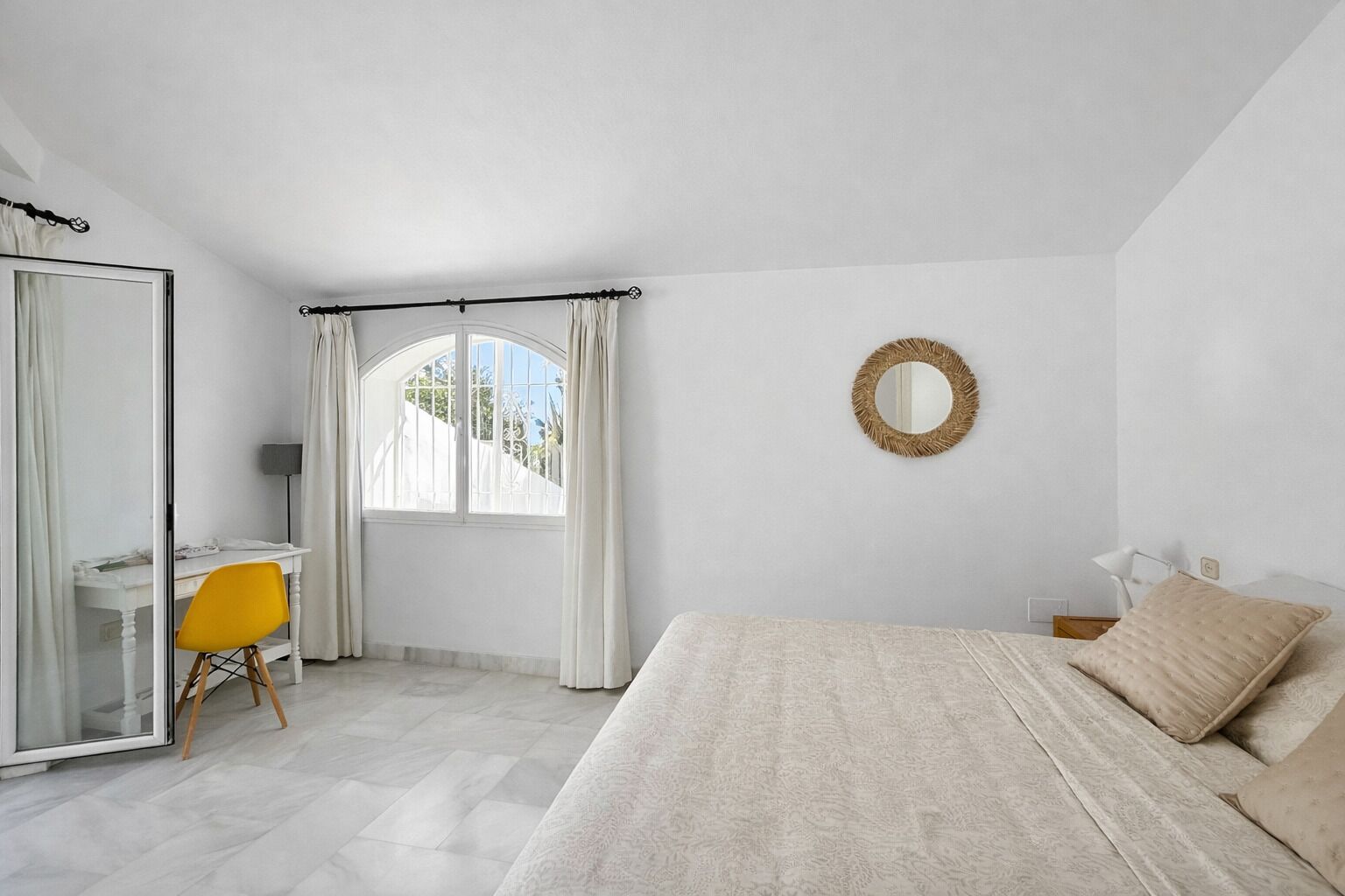 A bedroom with a bed, a desk, a chair, a mirror, and a window with curtains.
