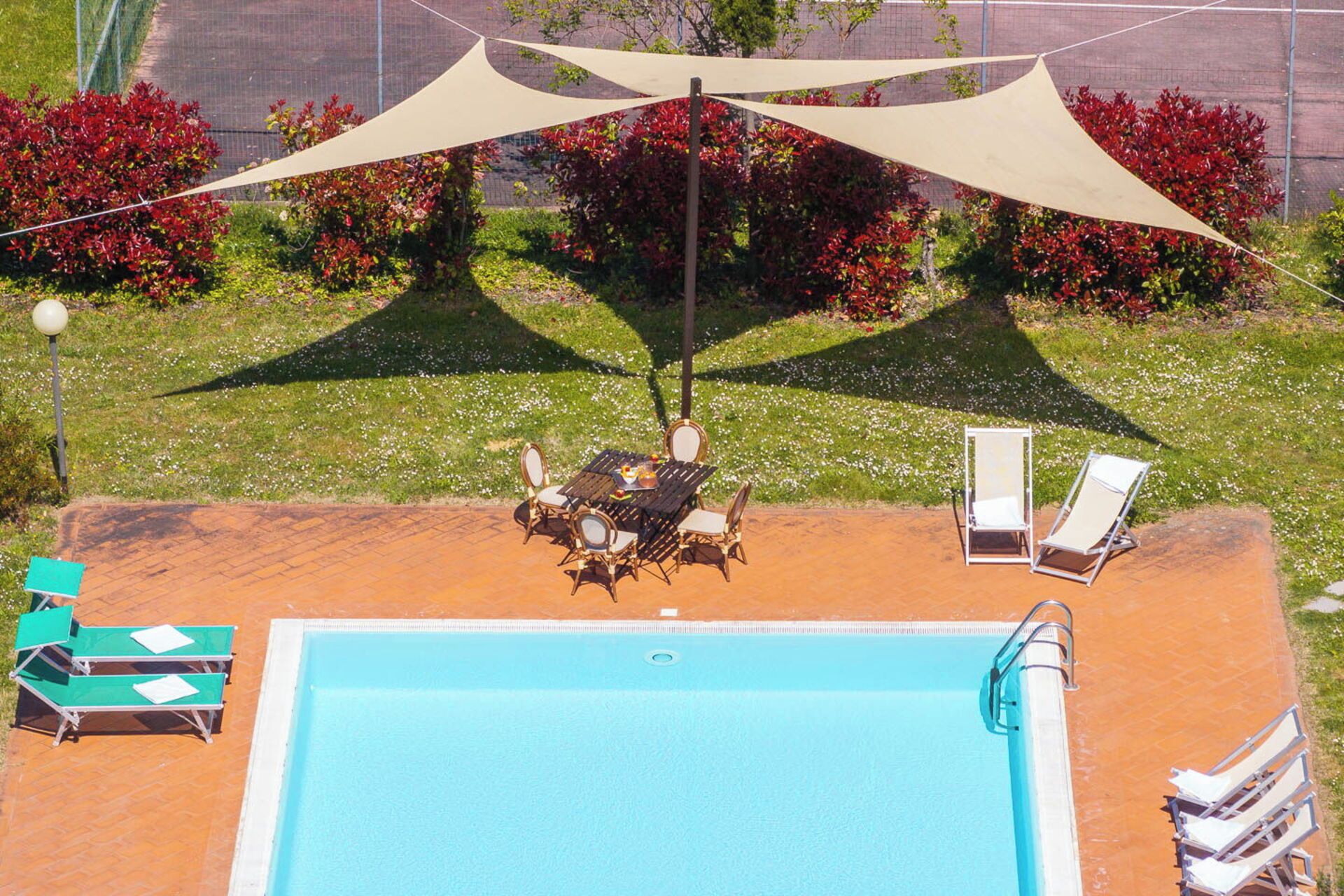 A swimming pool with a table and chairs under a shade sail, surrounded by greenery and red foliage.